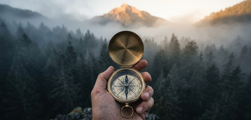 A hand holding a antique brushed-brass nautical compass over an open, fog-covered dense pine forest viewed from a high ridge A hand holding a antique brushed-brass nautical compass over an open, fog-covered dense pine forest viewed from a high ridge
