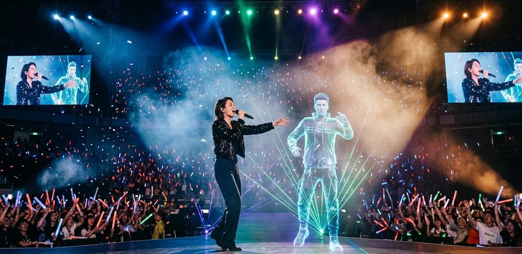 A real human female vocalist performing on stage alongside a holographic 3D male synthetic artist