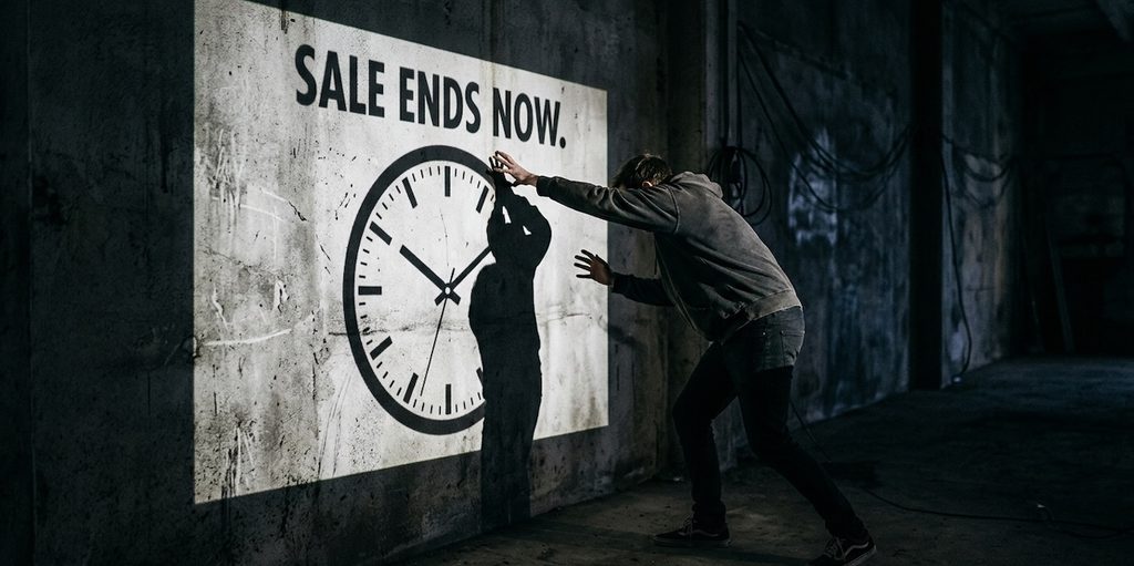 photo of a person trying to reach for the shadow of a clock cast onto a concrete wall by a projector displaying SALE ENDS NOW